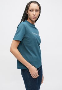 Tyrkysové bavlněné tričko s krátkými rukávy a bílým logem "Calvin Klein Jeans" na hrudi. Kulatý výstřih, volný střih.