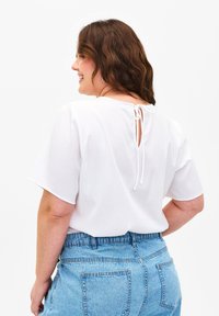 Top bianco a maniche corte con superficie texturizzata e chiusura a fiocco sul retro, abbinato a pantaloni shorts in denim blu a vita alta.