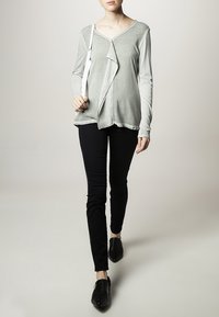Maglia grigia chiara a maniche lunghe con un design anteriore asimmetrico, abbinata a pantaloni neri aderenti e scarpe flat nere.