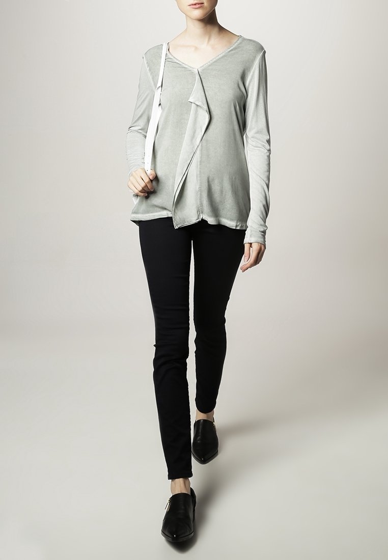 Maglia grigia chiara a maniche lunghe con un design anteriore asimmetrico, abbinata a pantaloni neri aderenti e scarpe flat nere.