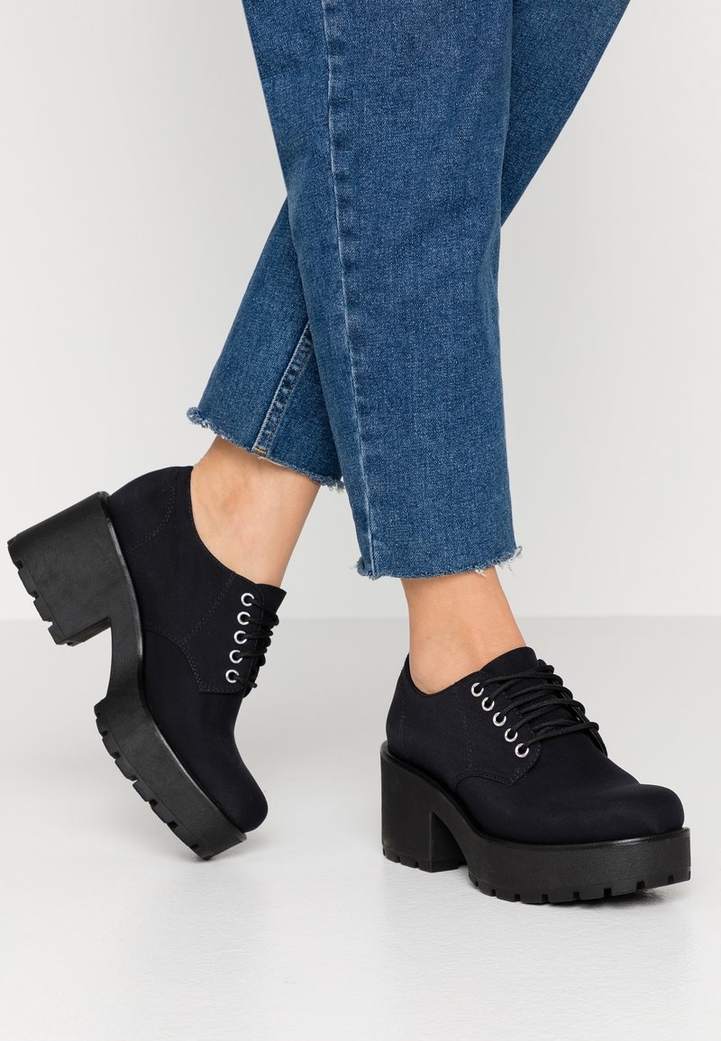 Chaussures à plateforme noires avec une tige en tissu lisse, bout rond et semelle épaisse en caoutchouc. Design à lacets avec œillets métalliques, portées avec un jean bleu.
