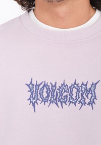 Felpa viola chiaro realizzata in tessuto morbido, con un logo grafico blu dai bordi frastagliati al centro. Collo a giro e bordo a costine.