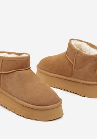 Bottes ankle en daim marron avec une semelle plate, doublure en fausse fourrure, bout arrondi et détails de couture près de la cheville.