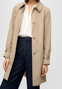 Kobieta w beżowym płaszczu typu trencz z guzikami, nosząca białą bluzkę i ciemnoniebieskie spodnie o wysokim stanie, stoi na tle w jednolitym kolorze.