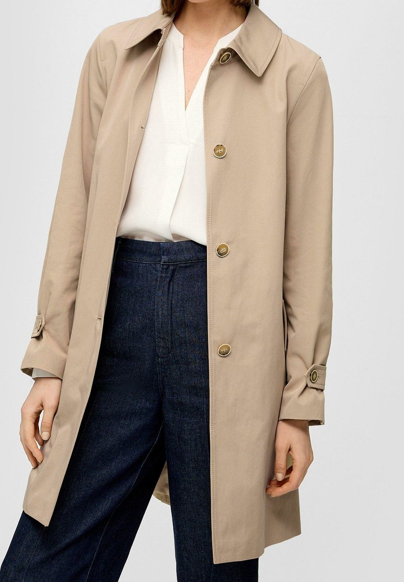 Kobieta w beżowym płaszczu typu trencz z guzikami, nosząca białą bluzkę i ciemnoniebieskie spodnie o wysokim stanie, stoi na tle w jednolitym kolorze.