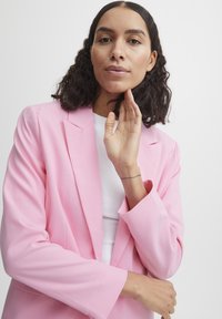 Light pink blazer with notched lapels, structured shoulders, and long sleeves. Worn over a white top, features smooth fabric and relaxed fit.