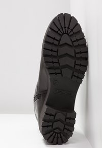 Botte noire avec une semelle en caoutchouc texturé présentant un motif de crampons et une légère élévation. Le talon est solide avec un branding sur le côté.