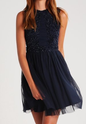 Ärmelloses marineblaues Kleid mit besticktem Oberteil und transparentem Tüllrock, getragen von einer Person mit langen braunen Haaren, vor einem schlichten Hintergrund.