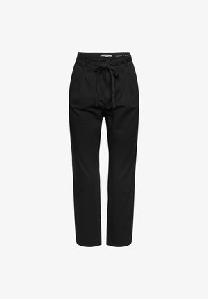 Persona que lleva una camisa blanca abotonada, pantalones negros, botas negras hasta el tobillo, sosteniendo una chaqueta burdeos doblada bajo el brazo.