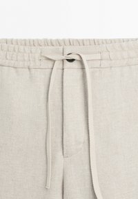 Close-up of beige pants waistband with elastic band and drawstring tied in a simple knot at the center front.