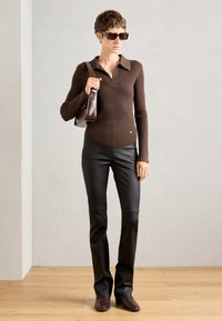 Rjava rebrasta dolgosleeve majica s ovratnikom, črne oprijete usnjene hlače, rjava torbica in temne sončne očala. Lesena tla v ozadju.