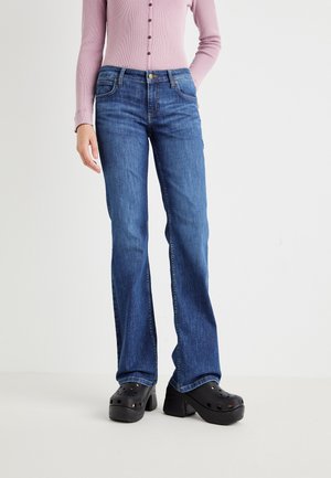 Jeans a zampa d'elefante blu con un lavaggio medio, materiale in denim robusto, design a cinque tasche e chiusura con bottone in vita, abbinati a zoccoli neri.