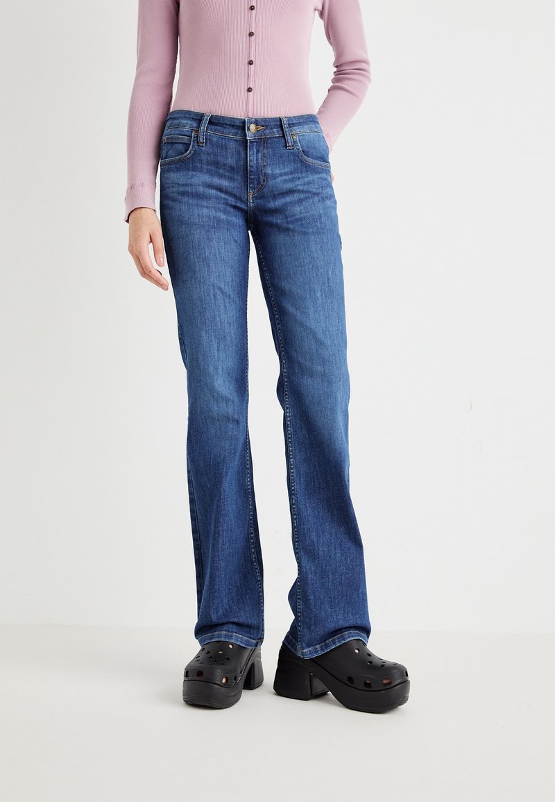 Jeans a zampa d'elefante blu con un lavaggio medio, materiale in denim robusto, design a cinque tasche e chiusura con bottone in vita, abbinati a zoccoli neri.