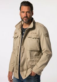 Giacca beige con design a colletto, due tasche sul petto e vita regolabile, realizzata in un tessuto testurizzato. Camicia nera a maniche lunghe sotto.