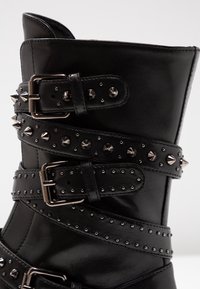 Czarny skórzany but z wieloma paskami, srebrnymi klamrami i metalowymi ćwiekami. Charakteryzuje się wyraźną formą i teksturowaną powierzchnią.