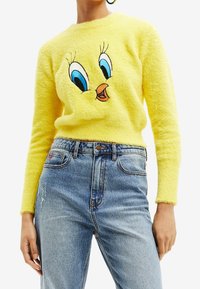 Suéter amarillo esponjoso con un personaje de dibujos animados bordado que tiene grandes ojos azules y un pico, combinado con jeans de tiro alto azules.