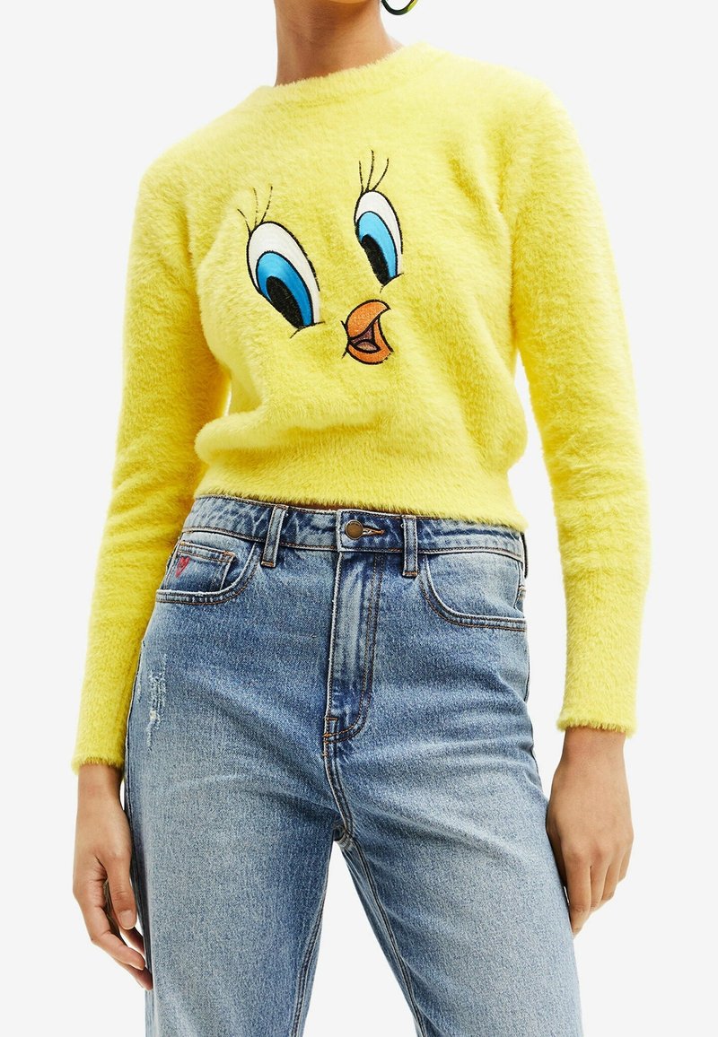 Suéter amarillo esponjoso con un personaje de dibujos animados bordado que tiene grandes ojos azules y un pico, combinado con jeans de tiro alto azules.