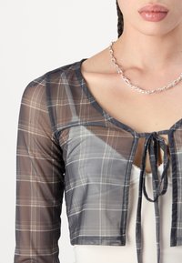 Graham kariertes Mesh-Crop-Top mit V-Ausschnitt, langen Ärmeln und einer Bindeschließe vorne. Verfügt über ein transparentes Material und kontrastierende Nähte.