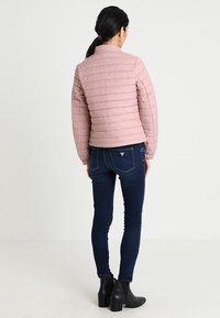 Chaqueta acolchada rosa con cuello alto, diseño acolchado y cierre de cremallera, combinada con jeans skinny de color azul oscuro y botas negras de tobillo.