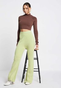 Bruine lange mouw cropped top gecombineerd met lichtgroene geribbelde flared broeken. Model draagt witte hakken. Eenvoudige, schone achtergrond.