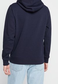 Tmavě modrá mikina s kapucí vyrobená z měkké látky, s kapucí na šňůrku, žebrovanými manžetami a lemem. Pohled zezadu ukazuje hladkou strukturu a volný střih.