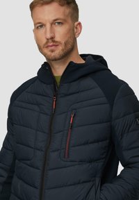 Navy pufferjack met een gestructureerd, geborduurd ontwerp, voorzien van een capuchon en contrasterende donkere mouwen. Inclusief een ritszak op de borst met rode accenten.