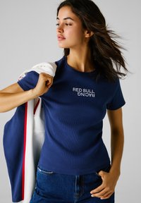 Camiseta azul marino de manga corta y con relieve, con "RED BULL RACING" bordado en blanco. Combinada con jeans de mezclilla oscura y una chaqueta ligera colgada del brazo.