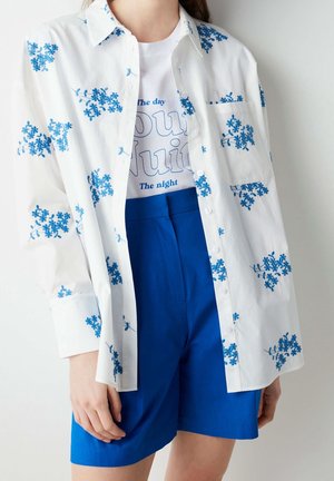 Person trägt ein weißes Hemd mit blauem Blumenmuster über einem weißen Grafik-T-Shirt und leuchtend blaue Shorts mit hoher Taille.