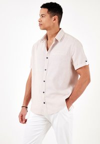 Camisa de manga corta beige con botones de tela texturizada, botones en color marino y un bolsillo en el pecho. Combinada con pantalones blancos.