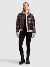 Gewatteerde jas in donker bordeauxrood met een capuchon gevoerd met schapenvacht, beschikt over een glanzende buitenkant, contrasterende crème binnenkant en zijzakken. Zwarte legging en combatlaarzen.