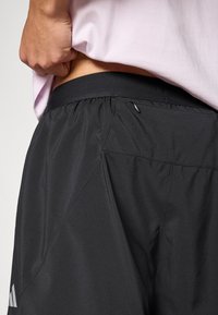 Fekete sportnadrág elasztikus derékpánttal, cipzáras zsebbel és sima tapintású anyagból. Az Adidas logó látható az oldalán.