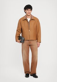 Chaqueta marrón de cremallero con cuello estampado, combinada con pantalones rectos marrón claro y zapatos negros, llevando un pequeño bolso negro.
