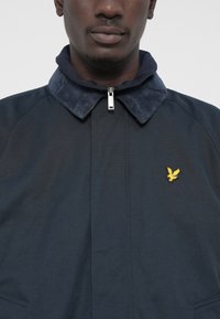 Navy jacket with a corduroy collar, featuring a front zipper and a small yellow emblem. Textured fabric and tailored design accents.