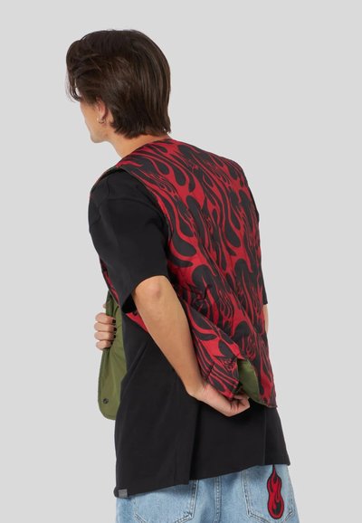 Gilet con motivo a fiamma rossa e nera sopra una maglietta nera, con accenti verdi visibili. Il gilet ha una texture liscia e una vestibilità ampia.
