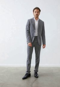 Completo grigio con giacca fitted e pantaloni, abbinato a una camicia bianca. Scarpe in pelle nera completano l'outfit. Tessuto liscio, vestibilità su misura.