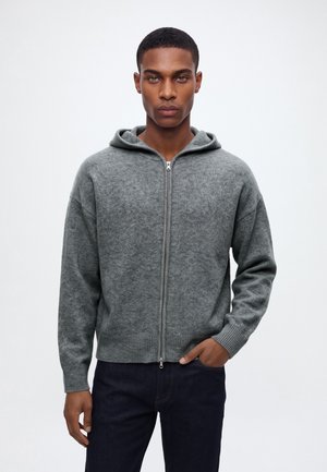 Jeune homme portant un sweat à capuche gris zippé avec un motif subtil et un pantalon foncé, debout avec une main dans la poche sur un fond uni.