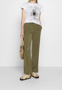 Camiseta blanca de algodón con una ilustración impresa, pantalones de pierna ancha de color oliva, sandalias beige de tipo deslizante y un bolso de paja tejido.