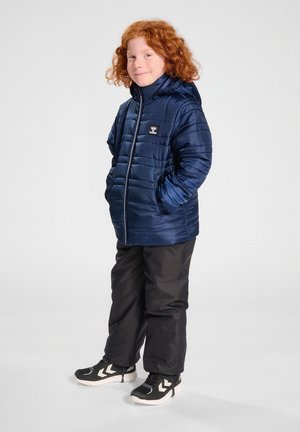 Barn med krøllet rødt hår iført en navyblå dynejakke, sorte bukser og sorte sportssko med hvide såler, står med hænderne i lommerne.