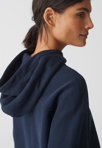 Felpa navy con una texture morbida, caratterizzata da un ampio cappuccio e scollatura aperta, mostrata di lato, evidenziando la caduta e i dettagli del tessuto.