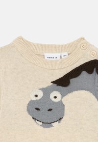 Beige stickad tröja med en grå drakansiktsdesign, med stora vita ögon och mörkbruna accenter, inklusive knappar på axeln.