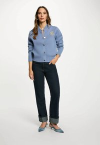 Cardigan lavorato a maglia di colore azzurro chiaro con colletto tondo, chiusura a bottoni, polsini a costine e un patch ricamato. Abbinato a jeans scuri, arrotolati sui polsini.
