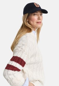 Frau mit langen blonden Haaren, die eine marineblaue Kappe und einen weißen Pullover mit Kabelstrickmuster sowie roten und weißen Streifen an den Ärmeln trägt.