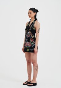 A young woman stands sideways, wearing a black dress with colorful floral and animal designs, paired with black platform sandals.