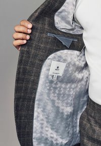 Maßgeschneiderte Blazer mit einer dunkelbraunen Karomuster-Außenfläche, silbernem geometrischem Musterfutter, Innentasche und angehängtem Markenschild innen.