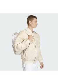 Chaqueta bomber beige claro con detalle de logo bordado, cierre de cremallera y puños acanalados, combinada con una mochila blanca con correas ajustables.