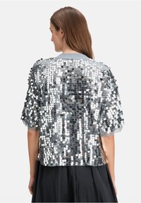 Silberner Paillettenjacke mit transparentem Rücken und kurzen Ärmeln. Hat einen runden Halsausschnitt und einen weichen grauen Saum. Strukturiert glänzende Oberfläche.