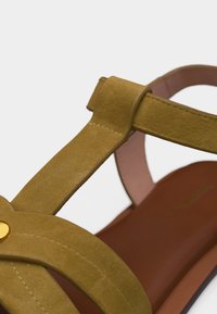 Κοντινή λήψη λουριού σανδαλιού από μουσταρδί κίτρινο σουέντ με χάλκινη μεταλλική διακόσμηση και καφέ δερμάτινη σόλα.