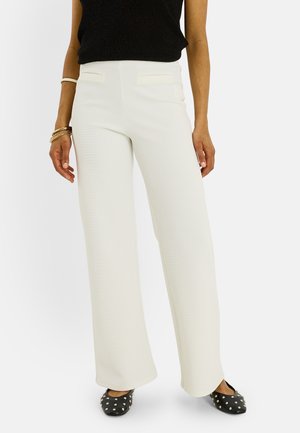 Mujer con pantalones blancos de talle alto y pernera ancha, top negro sin mangas, zapatos planos negros con tachuelas y brazaletes dorados en la muñeca izquierda.