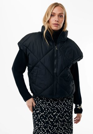 Frau trägt eine schwarze, kurzärmelige gesteppte Weste über einem schwarzen Langarmshirt und einem schwarzen Rock mit weißen Punkten, steht vor weißem Hintergrund.