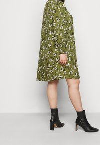 Abito floreale verde con maniche lunghe a sbuffo, taglio al ginocchio e stivaletti neri con tacco alto. Presenta piccoli fiori bianchi sparsi su tutto il vestito.